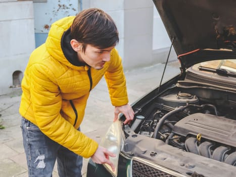 A young caucasian guy in a yellow jacket inspects an internal breakdown with an open hood of his car on a city street in front of an old house, close-up side view. The concept of engine repair, car repair, small, private, business, mechanic services.