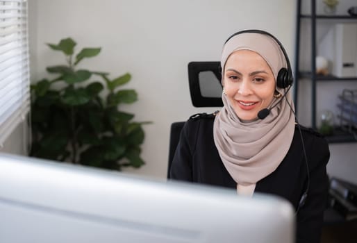 Call center worker, young Muslim woman wearing hijab, talking to customer on call phone on computer in customer service office.