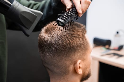 Stylist brushes man short hair using blowing dryer in barbershop closeup. Skilled barber does elegant hairstyle in beauty salon. Male haircut