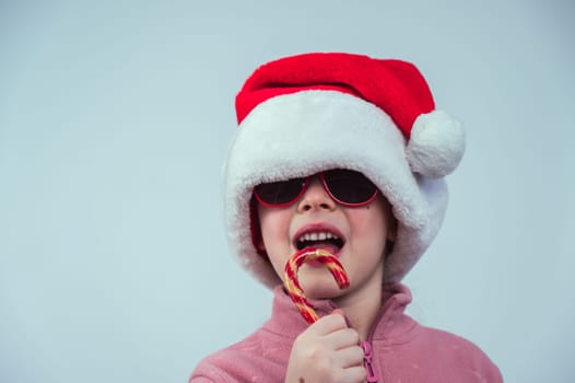 Portrait of a cute Caucasian girl in a Santa Claus hat and sunglasses eating a lollipop on a white background