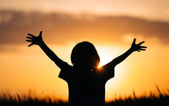 Silhouette of little boy - child kid with open hands in beautiful nature, sunset orange sky background. Freedom, happy childhood, summer holidays. High quality photo