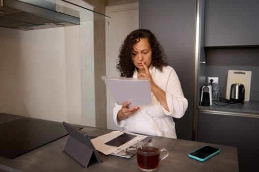 Young serious woman in white bathrobe, analyzing documents, doing paper work at home. Multi ethnic pretty brunette holding hands on lips while going through bills, looking worried