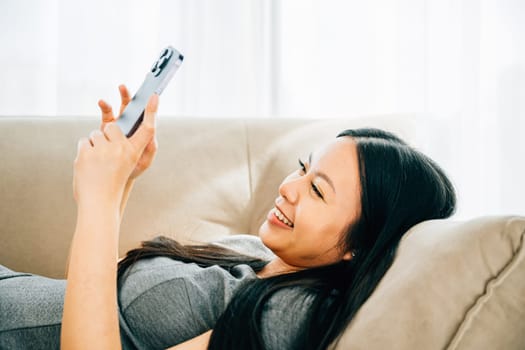 Asian woman on couch texts on smartphone smiling. Enjoying relaxation chatting and online shopping. Modern technology for communication and connection at home.