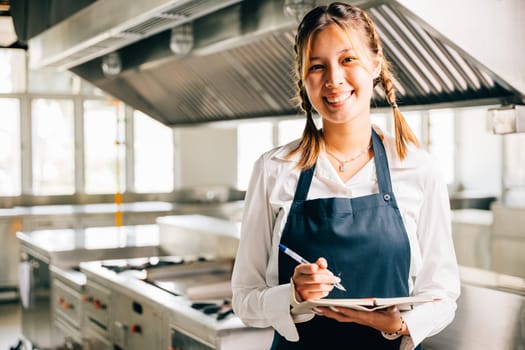 Portrait of a confident woman chef in commercial kitchen. Holds note book teaching with pen. Professional expertise explaining skills working with colleagues showing confidence. Foor education