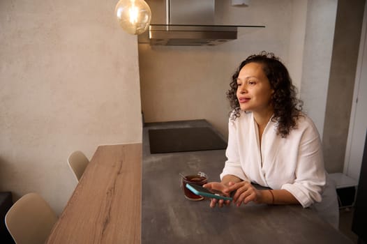 Attractive curly haired brunette woman in white bathrobe, holding mobile phone while standing at table in the home, dreamily looking away, texting message, scrolling newsfeed, online shopping
