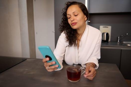 Attractive Latin American young woman in white bathrobe, using smartphone while drinking morning tea in kitchen at home. Relaxed positive lady reading text messages on her mobile phone and smiling