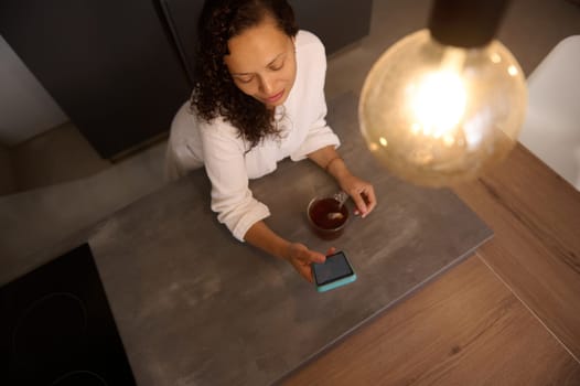 View from above of a multi ethnic beautiful young woman using her mobile phone while drinking a cup of tea in the kitchen at home. Mockup blank digital screen on smartphone. Copy advertising space
