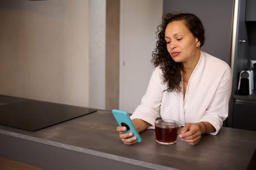 Chat in morning, news and breakfast at home. Happy young lady drinking tea and looking at smartphone, standing in modern minimalist home kitchen interior. Copy advertising space