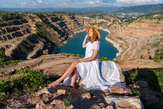 A woman in a white dress is sitting on a rock overlooking a body of water. The scene is serene and peaceful, with the woman enjoying the view and the calmness of the surroundings