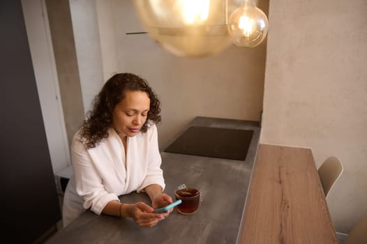 Happy Arab woman browsing social networks on smartphone and drinking tea in the kitchen at home. Gorgeous young Middle Eastern brunette using mobile phone for online communication. Free ad space