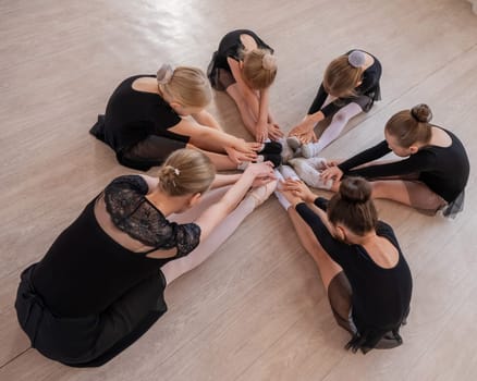 Caucasian woman and five little girls sit in a circle and do stretching at a ballet school