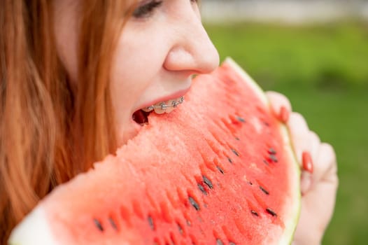 Close-up portrait of red-haired young woman with braces eating watermelon outdoors.