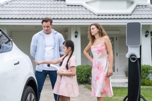 Happy little young girl learn about eco-friendly and energy sustainability as she help her family recharge electric vehicle from home EV charging station. EV car and modern family concept. Synchronos