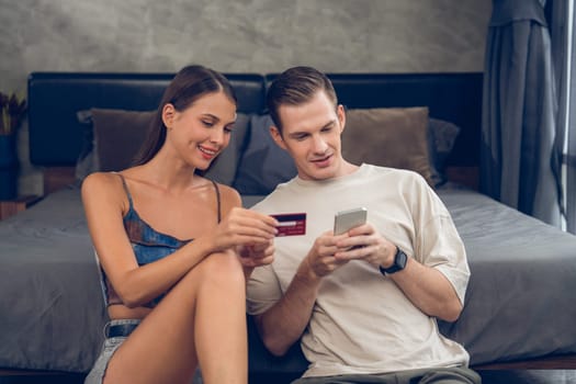 Young couple sit in the home bedroom using online payment app and digital wallet on smartphone to pay with credit card. E commerce shopping and modern purchasing via mobile internet. Unveiling