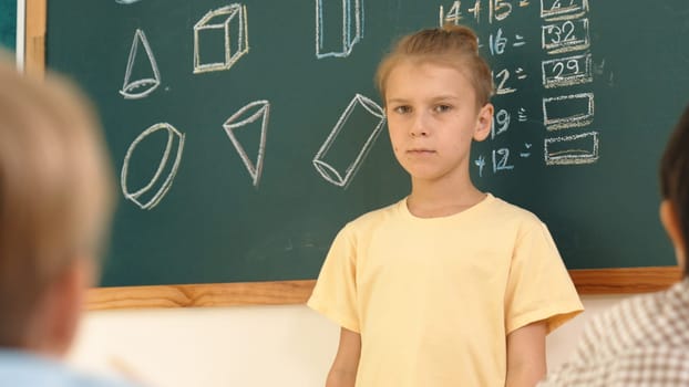 Caucasian boy talking and present idea while standing in front of blackboard with Math theory written. Diverse cute student sitting while looking at new student introduce himself. Education. Pedagogy.