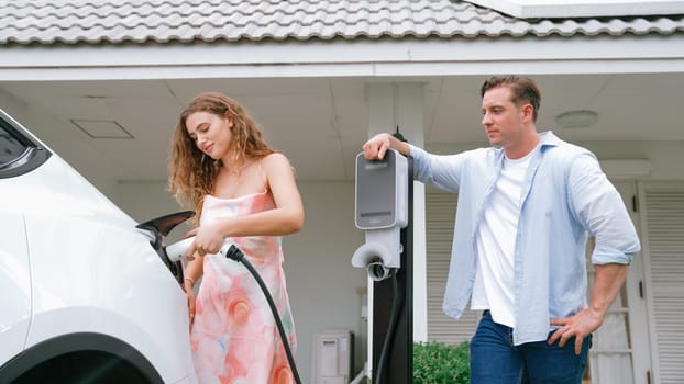 Happy and lovely couple with eco-friendly conscious recharging electric vehicle from EV charging station. EV car technology utilized as alternative transportation for future sustainability. Synchronos