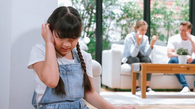 Stressed and unhappy young girl huddle in corner, cover her ears blocking sound of her parent arguing in background. Domestic violence at home and traumatic childhood develop to depression. Synchronos