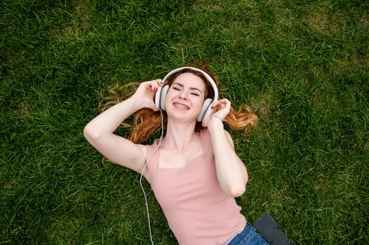 Top view of a young red-haired woman lying on the grass and listening to music on headphones