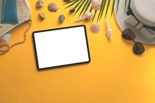 Digital tablet with blank screen, sunglasses, straw hat and seashells on yellow background.