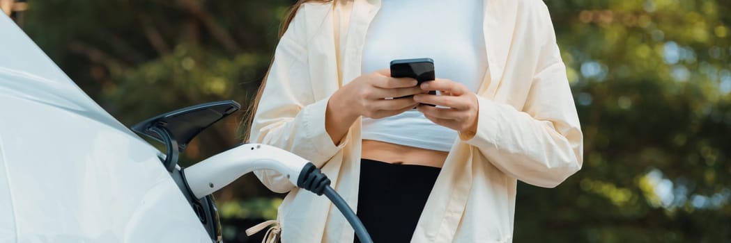 Young woman use smartphone to pay for electricity at public EV car charging station green city park. Modern environmental and sustainable urban lifestyle with EV vehicle. Panorama Expedient