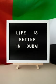 Message LIFE IS BETTER IN DUBAI on background of waving UAE flag made from silk. United Arab Emirates flag with concept of tourism and traveling. Inviting greeting card, advertisement. Dubai welcoming card