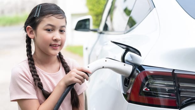 Happy little young girl learn about eco-friendly and energy sustainability as she recharge electric vehicle from home EV charging station. EV car and sustainable future generation. Panorama Synchronos