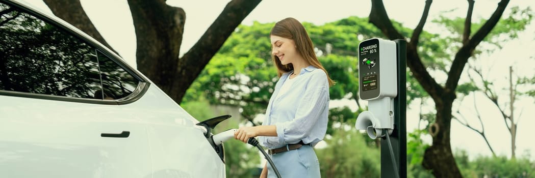 Young woman recharge EV electric vehicle's battery from EV charging station in outdoor green city park scenic. Eco friendly urban transport and commute with eco friendly EV car travel. Panorama Exalt