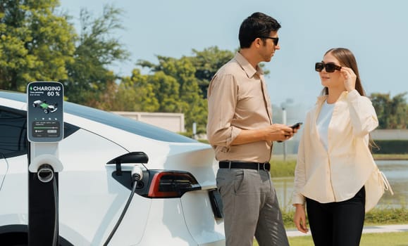 Young couple use smartphone to pay for electricity at public EV car charging station green city park. Modern environmental and sustainable urban lifestyle with EV vehicle. Expedient