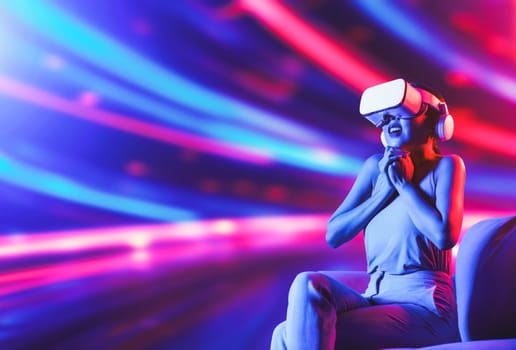 Smart female sitting on sofa surrounded by neon light wearing VR headset connecting metaverse, future cyberspace community technology. Elegant woman excited and emotionally watch movie. Hallucination.