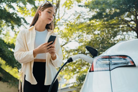 Young woman use smartphone to pay for electricity at public EV car charging station green city park. Modern environmental and sustainable urban lifestyle with EV vehicle. Expedient