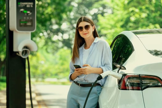 Young woman using smartphone online banking application to pay for electric car battery charging from EV charging station during vacation holiday road trip at national park or summer forest. Exalt