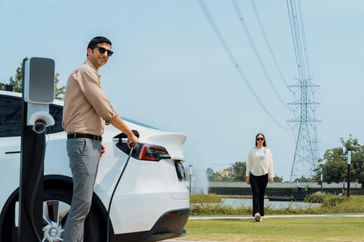 Young couple recharge EV car battery at charging station connected to power grid tower electrical industrial facility as electrical industry for eco friendly vehicle utilization. Expedient