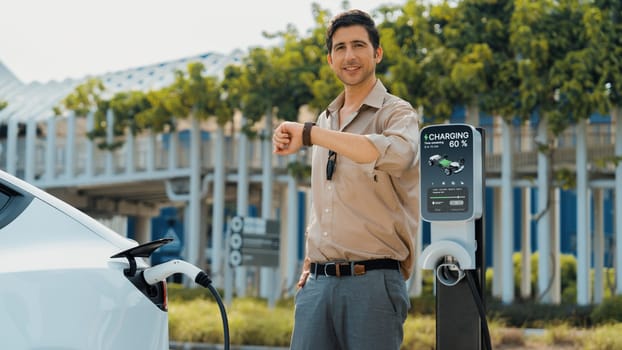 Young man checking time on smartwatch while EV charger to recharging battery from charging station in parking lot. Rechargeable EV car for sustainable eco friendly urban travel. Panorama Expedient