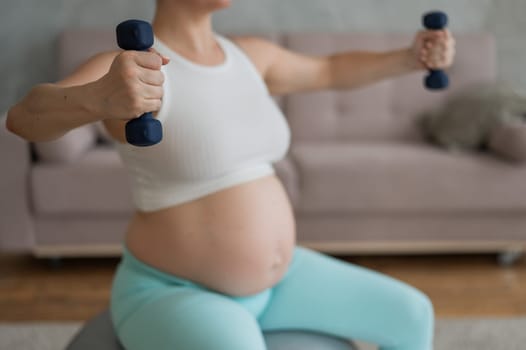 Pregnant woman doing exercises with dumbbells while sitting on a fitness ball at home. Close-up of a pregnant belly