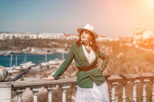 Woman walks around the city, lifestyle. Happy woman in a green jacket, white skirt and hat is sitting on a white fence with balusters overlooking the sea bay and the city