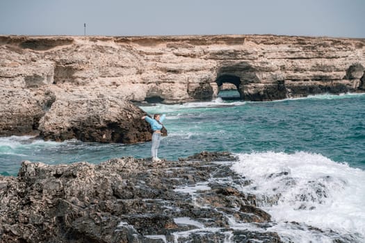 A woman in a blue jacket stands on a rock above a cliff above the sea and looks at the raging ocean. Girl traveler rests, thinks, dreams, enjoys nature. Peace and calm landscape, windy weather