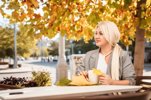 A blonde woman sits at a table with a cup of coffee and a leaf on it. The scene is set in a city with a tree in the background