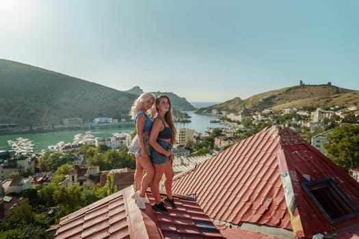 women standing on rooftop, enjoys town view and sea mountains. Peaceful rooftop relaxation. Below her, there is a town with several boats visible in the water. Rooftop vantage point