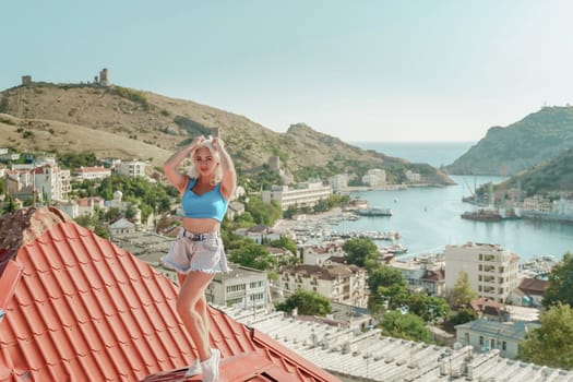 Woman standing on rooftop, enjoys town view and sea mountains. Peaceful rooftop relaxation. Below her, there is a town with several boats visible in the water. Rooftop vantage point