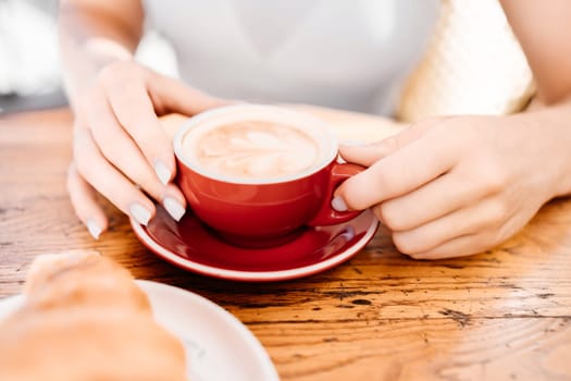 A woman is holding a red coffee cup with a white saucer. The woman is sitting at a table with a plate of croissants in front of her