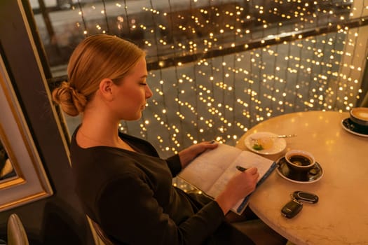 A woman is sitting at a table with a notebook and a cup of coffee. She is writing in her notebook while looking out the window. The scene suggests a moment of relaxation and contemplation