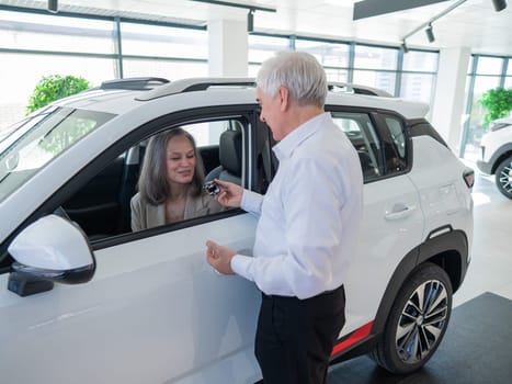 Mature Caucasian woman sits in a new car, an elderly man gives her the keys