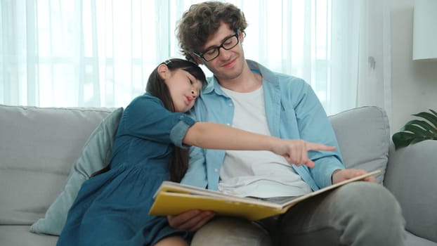 Happy daughter lean on father shoulder while listening story telling. Young asian schoolgirl looking ar dad while reading story book together and sitting at sofa. Family recreation concept. Pedagogy.