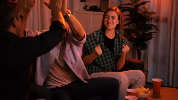 Family and colleague sitting at dark living room with red filter while cheering football team together. Caucasian people enjoy watching tv and giving high five to celebrate winning team. Convocation.