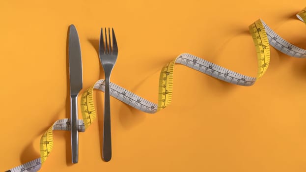 Fork and knife wrapped with measuring tape on yellow background. Weight loss and healthy concept.