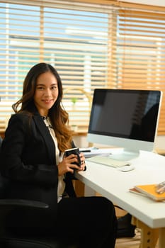 Portrait of successful businesswoman with cup of coffee sitting at desk and smiling at camera.