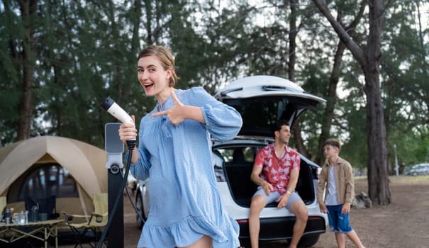 Outdoor adventure and family vacation camping at sea travel by eco friendly car. Cheerful woman or mother holding, pointing EV charger point with playful and happiness posture in campsite. Perpetual