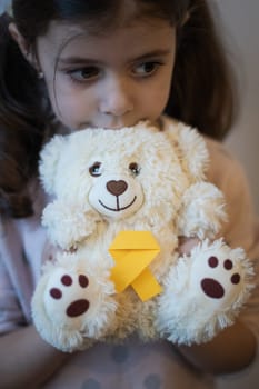 Beautiful little caucasian brunette female Holds in her hands holds a teddy bear with a yellow ribbon tie thoughtfully looking to the side on a white background,close-up side view with depth of field. World childhood cancer day concept.