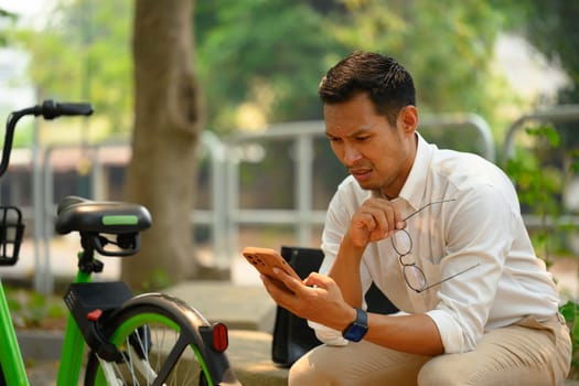 Serious millennial businessman holding eyeglasses reading message from mobile phone.