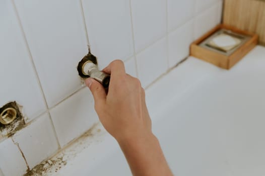 One young Caucasian unrecognizable guy takes out a faucet nut from a hole in a white tiled wall while standing in the bathroom at home, close-up side view.Step by step.